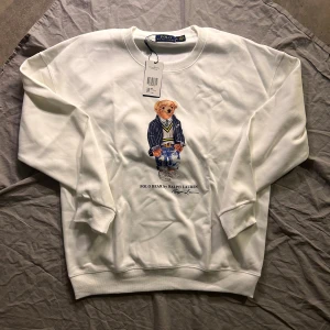 Vit sweatshirt Polo Bear Ralph Lauren  - Vit sweatshirt från Polo Ralph Lauren i storlek XL med ikoniskt Polo Bear-tryck på bröstet. Tröjan har rund hals, ribbade muddar och är tillverkad i mjuk bomull. Perfekt för dig som gillar klassisk streetstyle med en twist.