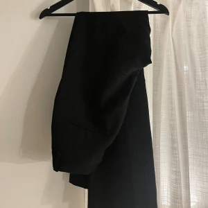 Svart, högmidjad kostymbyxa - Snygga svarta kostymbyxor med utsvängd passform och diskreta fickor. Perfekt för dig som vill ha en stilren och tidlös look. Materialet känns mjukt och har en lätt glans, vilket ger en elegant känsla. 