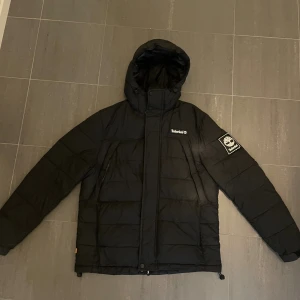 Svart pufferjacka från Timberland - Svart pufferjacka från Timberland med huva och snygga logodetaljer på bröstet och ärmen. Jackan har dragkedja framtill, quiltad design och två sidofickor och 2 bröstfickor med dragkedja. Perfekt för kalla dagar och har en clean, urban look som passar streetwear. Inga skador.