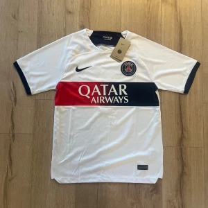 PSG Mbappé vit matchtröja Nike  - Paris Saint-Germain fotbollströja med Mbappé och nummer 7 på ryggen. Vit med breda mörkblå och röda ränder över bröstet, PSG-emblem och Nike-logga. Tillverkad i lätt polyester, kortärmad och med Qatar Airways tryck framtill.