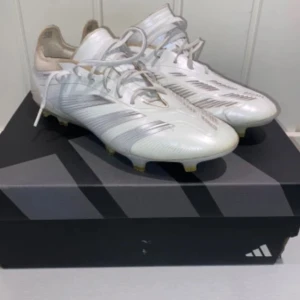 adidas Predator Elite FG Pearlized - Säljer ett par snygga vita fotbollsskor från Adidas med silverfärgade detaljer och snörning. Skorna har en låg profil och platt sula med dobbar för gräsplan. Ovandelen är i syntetmaterial och insidan har perforerat tyg för ventilation. Perfekt för dig som vill ha stil på planen. Använda 5-7 gånger!
