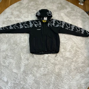 Svart vindjacka från Corteiz - Svart vindjacka från Cortez Guerillaz med huva och coolt grått graffiti-mönster på axlar och ärmar. Jackan har broderad ELITEWORK-logga på bröstet, dragkedja framtill och snörning i nederkant. Perfekt för dig som gillar streetwear och unik stil. Använd ett fåtal gånger! Storlek M men är rätt kort i längden så passar S perfekt också! 