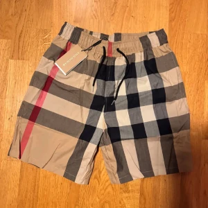 Rutig shorts från Burberry - Snygga shorts från Burberry med klassiskt rutigt mönster i beige, svart, vitt och rött. De har elastisk midja med snörning och är perfekta för sommaren. Materialet känns lätt och bekvämt.