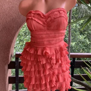 Axelbandslös korallrosa volangklänning - Säljer en axelbandslös kort klänning i korallrosa med flera lager volanger och plisserade detaljer. Klänningen har en hjärtformad överdel och är perfekt för dig som gillar att sticka ut med färg och struktur. Materialet känns lätt och luftigt.