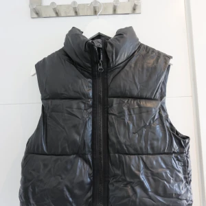 Svart dunväst med hög krage - Snygg svart dunväst med glansig finish och hög krage. Västen har en rejäl dragkedja framtill och quiltad design som ger en cool och modern look. Perfekt att slänga över en hoodie för extra värme under kyliga dagar.