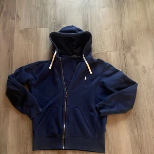 Marinblå hoodie från Polo Ralph Lauren - Snygg marinblå hoodie från Polo Ralph Lauren med vit broderad logga på bröstet. Tröjan har dragkedja, huva med vita snören och två stora fickor framtill. Perfekt för en chill och stilren look.