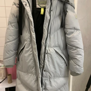 Ljusgrå lång pufferjacka Amisu - Säljer en ljusgrå lång pufferjacka från Amisu Essential Outerwear. Jackan har huva, svarta detaljer, dragkedja och praktiska fickor. Insidan har en cool neonfärgad zip och snygga etiketter. Perfekt för kalla dagar och riktigt stilren.