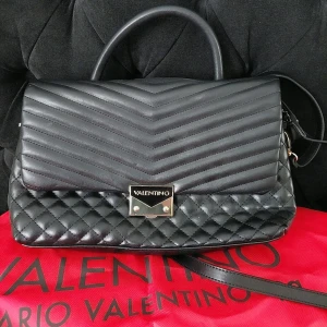 Svart quiltad handväska Valentino - Snygg svart handväska från Valentino med quiltad design och stilrena detaljer. Väskan har både handtag och axelrem, samt flera fack med dragkedja inuti för smart organisering. Materialet är skinn och väskan har en metallplatta med logga framtill.