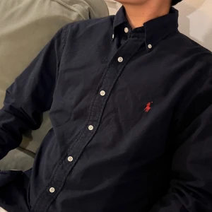 Mörkblå skjorta från Ralph Lauren - Snygg mörkblå skjorta från Ralph Lauren med klassisk button-down krage och vita knappar. På bröstet finns det ikoniska röda Polo-broderiet. Skjortan är långärmad och tillverkad i mjuk bomull, perfekt för en clean och stilren look.