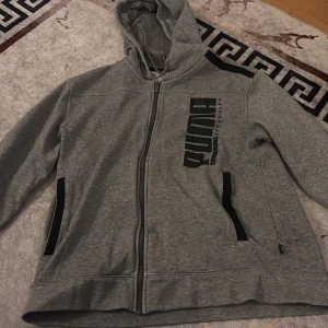 Grå hoodie från Puma med dragkedja - Grå hoodie från Puma med svart tryck på bröstet och svarta detaljer vid fickorna. Tröjan har huva med snörning, lång ärm och hel dragkedja framtill. Perfekt för chill eller träning, mjukt material och sportig look.