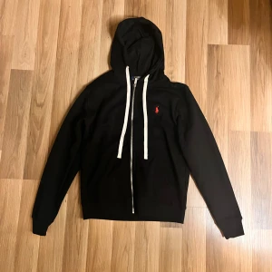 Svart hoodie från Polo Ralph Lauren - Svart hoodie från Polo Ralph Lauren med vit snörning och dragkedja framtill. Klassisk röd broderad logga på bröstet. Tröjan har huva, långa ärmar och är tillverkad i mjuk bomull. Perfekt för chill dagar och enkel att matcha med jeans eller joggers.