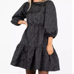 Svart blommig kortklänning med trekvarts puffärm - SKITSNYGG nyprsklänning med lyxigt tyg perfekt till nyår från NeoNoir. Aldrig använd och helt slutsåld. Perfekt till ett par nylon och höga stövlar eller klackar. Trekvartsärmar som ger plats för fina armband och går ovanför knäna med ett subtilt blom mönster för extra dyr känsla