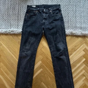 Levi's 501 svarta jeans W30 L32 - Svarta Levi's 501 jeans med klassisk rak passform och fem fickor. Jeansen har en snygg tvättad look, knappgylf och den ikoniska läderpatchen bak i midjan. Perfekt för dig som gillar tidlös stil och vill ha ett par jeans som funkar till allt.