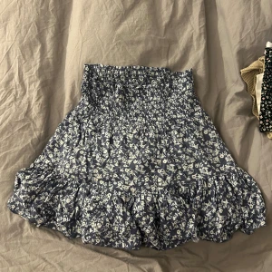 Blommig blå volangkjol från lager 157 - Supersöt kort kjol från lager 157 i blått med vitt blommönster. Kjolen har smock i midjan för skön passform och en volangkant längst ner som ger extra flöde. Perfekt för dig som gillar en feminin och chill stil. Materialet är mjukt och lätt bomull.