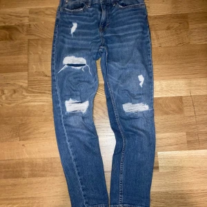 Blå slim fit jeans med slitningar - Snygga blå slim fit jeans från &Denim med flera slitningar och hål på benen för en trendig look. Klassisk femficksmodell med knapp och dragkedja. Tillverkade i denim med coola detaljer och passform som sitter tajt men bekvämt.