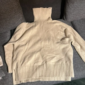 Beige stickad polotröja med ribbade detaljer - Beige stickad polotröja med hög, ribbad krage och breda ribbade muddar vid nederkant och ärmslut. Tröjan har en loose fit och är perfekt för lager-på-lager under kyliga dagar. Enkel och stilren design som passar till allt.