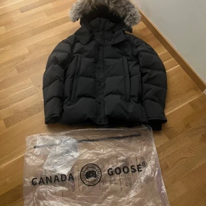 Canada Goose Wyndham  - Säljer en svart, riktigt fet dunjacka från Canada Goose med fluffig päls på huvan. Jackan har en klassisk puffer-look, snygga detaljer med både dragkedja och knappar, samt loggan på ärmen. Perfekt för kalla vinterdagar och har en schysst oversized vibe.