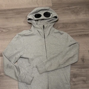 Grå c.p kompany zip tröja  - Säljer en grå c.p kompany zip tröja med goggles på luvan i svart. Tröjan har en bra passform och är perfekt för en klassisk stilig stil. Tröjan är köpt på NK men saknar tyvärr kvitto o boxen. Pris ej skrivet i sten