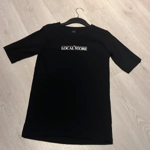 Svart t-shirt från HOOK Local Store - Svart t-shirt från HOOK med vit text 'THE LOCAL STORE' på bröstet. Klassisk rak modell med korta ärmar och rund hals. Tillverkad i 100% bomull för skön och mjuk känsla. Perfekt för dig som gillar enkel och stilren streetwear. Den har ett liten hål i nacken som ni kan se på bild 3. Det hålet är så litet att de inte syns