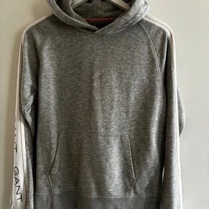 Grå hoodie från GANT med logga - Grå hoodie från GANT med vit logga längs ärmarna och klassisk känguruficka. Tröjan har huva och ribbade muddar. Perfekt för chill dagar och snygg till jeans eller joggers.XS