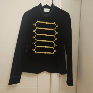 Svart kavaj i sammet från Neo Noir - Svart rock från Neo Noir i sammet med guldiga dekorativa snörningar och knappar framtill. Jackan har lång ärm och snygga broderade detaljer vid kragen och ärmslut. Stilren och unik design som sticker ut.