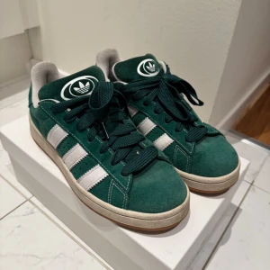 Adidas Campus gröna sneakers - Snygga Adidas Campus sneakers i mörkgrön mocka med klassiska vita ränder och logga på plösen. Skorna har rund tå, platt sula och snörning. Perfekta för dig som gillar retrostil och vill sticka ut med färg. Passar till både jeans och shorts.
