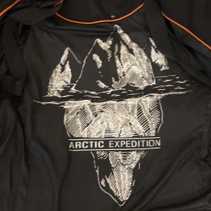 Svart lång pufferjacka Hollies 38 - Svart lång pufferjacka från Hollies Stockholm med coolt Arctic Expedition-tryck på insidan. Jackan har huva, flera dragkedjor, snygga detaljer och orange piping på insidan. Perfekt för kalla dagar och har en modern look med emblem på ärmen.
