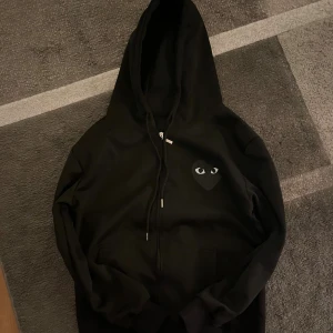 Svart hoodie med hjärta och ögon - Svart hoodie med dragkedja och huva. På bröstet och ryggen finns ett stort svart hjärta med vita ögon, vilket ger en unik och cool look. Tröjan har långa ärmar och fickor framtill. Perfekt för dig som gillar streetwear och vill sticka ut.