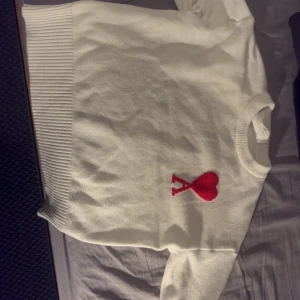 Vit stickad tröja från Ami med hjärta - Vit stickad tröja från Ami med ribbade muddar och rund hals. På bröstet finns ett rött hjärta med bokstaven A, vilket ger en unik och snygg detalj. Perfekt för dig som gillar stilrena plagg med en twist.