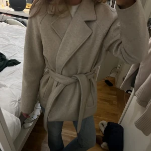 Beige kappa med bälte och krage - Säljer en beige kappa med bred krage och knytbälte i midjan. Jackan har en stilren och klassisk look med långa ärmar och mjukt material, perfekt för dig som gillar neutrala färger och enkel design.