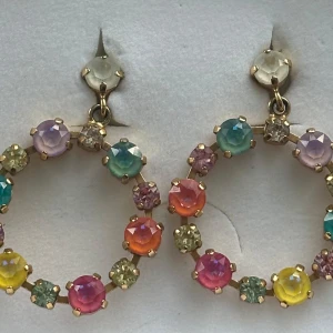 Färgglada cirkelörhängen med stenar - Säljer ett par stora cirkelörhängen med gnistrande stenar i flera färger som rosa, gul, blå, grön, lila och orange. Örhängena har guldfärgade detaljer och ger en riktigt färgstark och lekfull vibe. Perfekta för dig som vill sticka ut och addera färg till din look.