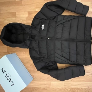 Svart pufferjacka från The North Face - Svart pufferjacka från The North Face i storlek M. Jackan har huva, dragkedja framtill och ett stilrent, quiltat mönster. Perfekt för kalla dagar och har en clean look med logga på bröstet. Materialet är syntet och jackan är långärmad.