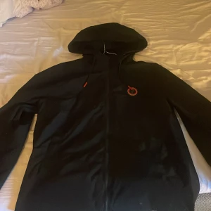 Svart vindjacka från Trapstar - Svart vindjacka från Trapstar med huva och dragkedja framtill. Jackan har röda detaljer på snörningen och ett Trapstar-tryck i rött på ryggen. Snygg och stilren design som passar perfekt till streetwear. 