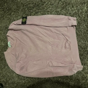 Ljusrosa Stone Island sweatshirt - Säljer en ljusrosa sweatshirt från Stone Island med den klassiska logotypen på vänster ärm. Tröjan har rund hals, ribbade muddar och är tillverkad i mjuk bomull. Perfekt för dig som gillar streetwear och vill ha något stilrent med ikonisk badge.