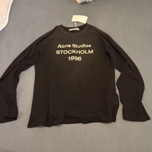 Svart långärmad tröja Acne Studios - Svart långärmad tröja från Acne Studios med tryck i vitt på bröstet där det står 'Acne Studios Stockholm 1996'. Tröjan har rund hals och är tillverkad i mjuk bomull. Perfekt för dig som gillar stilrena och trendiga plagg med tydlig logga.