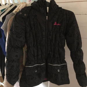 Svart pufferjacka från Desigual - Svart pufferjacka från Desigual med coolt paisleymönster och broderad rosa logga på bröstet. Jackan har huva, dragkedja framtill, fickor med knappar och en färgglad patch baktill. Insidan är fodrad i stark rosa. Perfekt för dig som gillar unika detaljer.