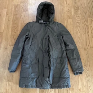 Grå parkajacka från Woolrich - Säljer en grå parkajacka från Woolrich med huva och stora fickor framtill. Jackan har både dragkedja och knappar, samt ribbade muddar vid ärmslut. Perfekt för kalla dagar och har en stilren, längre passform.