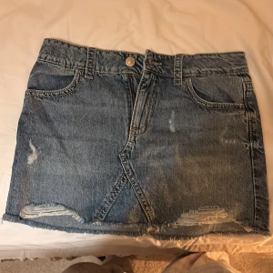 Blå jeanskjol från Gina Tricot - Säljer en blå jeanskjol från Gina Tricot i klassisk denim med slitna detaljer och rå kant nedtill. Kjolen har fem fickor, knapp och dragkedja framtill samt snygga bakfickor med knapp. Perfekt för dig som gillar en chill och trendig look.