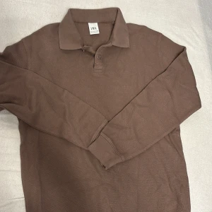 Brun pikétröja från Zara - Säljer en brun långärmad pikétröja från Zara i storlek S. Tröjan har klassisk krage och knäppning framtill, tillverkad i ett mjukt och bekvämt material. Perfekt för en chill och stilren look.