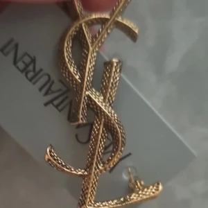  guldigt brosch med logotyp - Stilren brosch med den ikoniska i guldtonad metall. Bokstäverna har en snygg texturerad yta som ger extra lyxig känsla. Perfekt statement piece för dig som gillar exklusiva accessoarer.