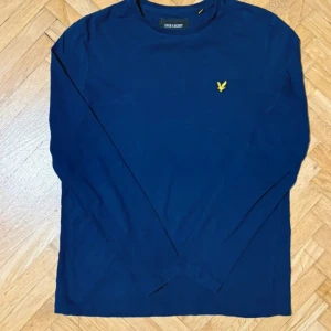Marinblå långärmad tröja Lyle & Scott - Snygg marinblå långärmad tröja från Lyle & Scott med gul logga broderad på bröstet. Klassisk rund hals och mjukt bomullsmaterial som känns skönt mot huden. Perfekt för dig som gillar stilrena och enkla plagg med en liten detalj.