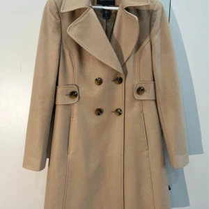 Beige kappa med stora knappar - Snygg beige kappa från Zara med dubbelknäppning och bred krage. Jackan har långa ärmar och dekorativa knappar vid midjan. Perfekt för dig som gillar klassisk stil med modern touch.