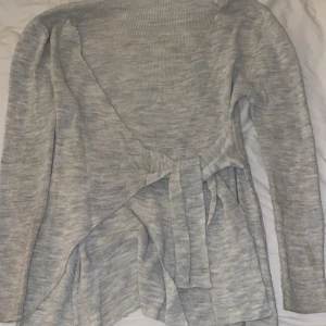 Grå omlott kofta med knytband - Snygg grå ribbad omlottkofta med långa ärmar och knytband i midjan. Koftan har en mjuk och stretchig känsla, perfekt för lager-på-lager. Den har en stilren look och passar dig som gillar enkel men trendig design.