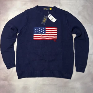 Marinblå stickad tröja Polo Ralph Lauren - Snygg marinblå stickad tröja från Polo Ralph Lauren med amerikansk flagga broderad på bröstet och RL-detalj i rött. Tröjan har rund hals och långa ärmar med ribbade muddar. Perfekt för dig som gillar klassisk stil med en twist.