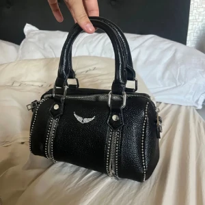 Svart handväska med silverdetaljer - Cool svart handväska i skinn med två rejäla handtag och silverfärgad logga framtill. Väskan har grova dragkedjor och nitar längs kanterna som ger en edgy vibe. Perfekt storlek för det viktigaste och snygg att bära både i handen och på armen.