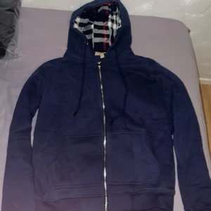 Marinblå Burberry hoodie - Snygg marinblå hoodie från Burberry i storlek XL, men sitter som L. Säljer åt min bror som har XL men tröjan satt mer som L för honom. Tröjan har dragkedja, huva med klassiskt rutigt Burberry-foder och broderad logga på bröstet. Tillverkad i mjuk bomull med praktiska fickor framtill och snörning i huvan. Perfekt för dig som gillar stilren design med lyxig touch. Helt ny och äkta inga defekter.
