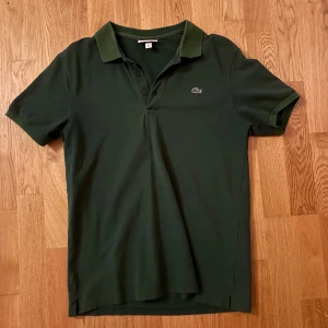 Grön pikétröja från Lacoste - Snygg mörkgrön pikétröja från Lacoste med klassisk krage och korta ärmar. Tröjan har knappar framtill och den ikoniska krokodilloggan på bröstet. Perfekt för dig som gillar stilrena och sportiga plagg.