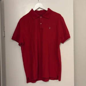 Röd pikétröja från Boomerang L - Snygg röd pikétröja från Boomerang i storlek L. Klassisk krage, korta ärmar och två knappar framtill. Tröjan har ett litet broderat boomerangmärke på bröstet och är gjord i mjuk bomull. Perfekt för dig som gillar stilrena och sportiga plagg.