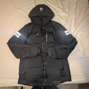 Svart Sail Racing dunjacka med huva - Säljer en svart dunjacka från Sail Racing, modell Antarctica Expedition. Jackan har huva, reflekterande detaljer på ärmarna och logga på bröstet. Flera praktiska fickor och dragkedja framtill. Perfekt för kalla dagar och riktigt snygg streetstil.