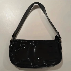 Svart axelväska i blankt material - Snygg svart axelväska med glansig finish och enkel design. Väskan har en bred axelrem och stängs med dragkedja upptill. Perfekt storlek för det viktigaste och ger en cool vibe till din outfit.